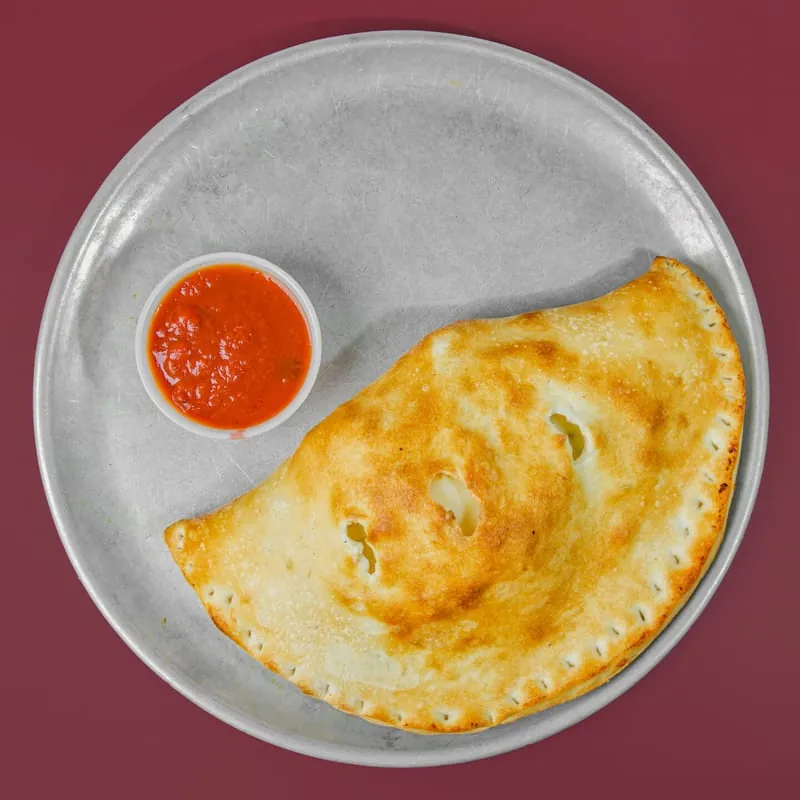 Calzone on a pizza pan with a cup of marinara